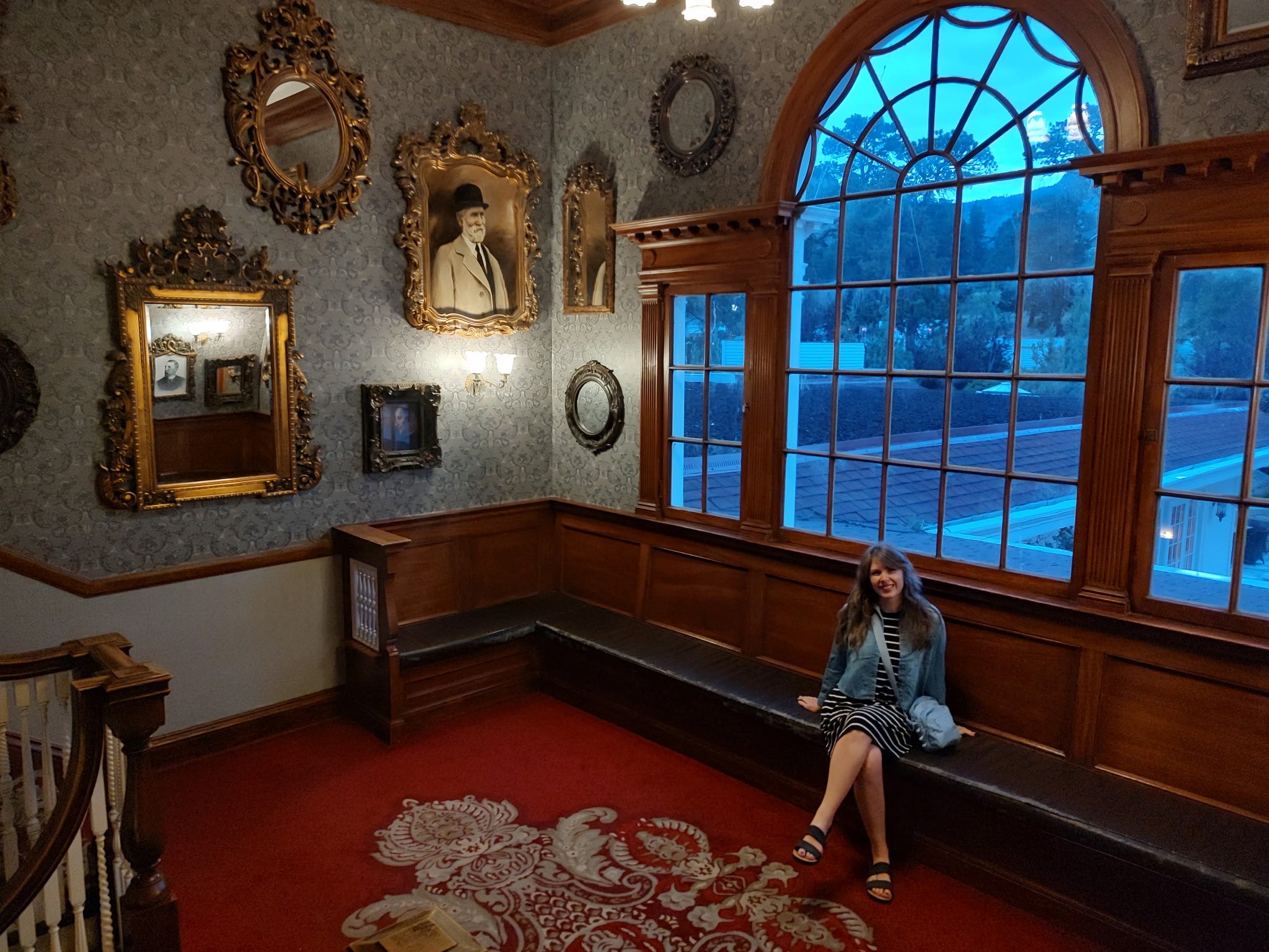A woman in a black dress with white stripes sits on a bench at the top of a staircase. There are gold mirrors on the wall